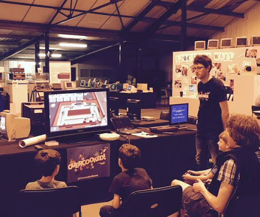 A group of children watching a gameplay demonstration of the cooperative cooking game Overcooked on a television screen, with a player interacting with the game and various gaming equipment visible in the background.