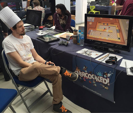 A person wearing a chef's hat sitting at a table playing the video game Overcooked on a monitor, with gaming equipment and people in the background.